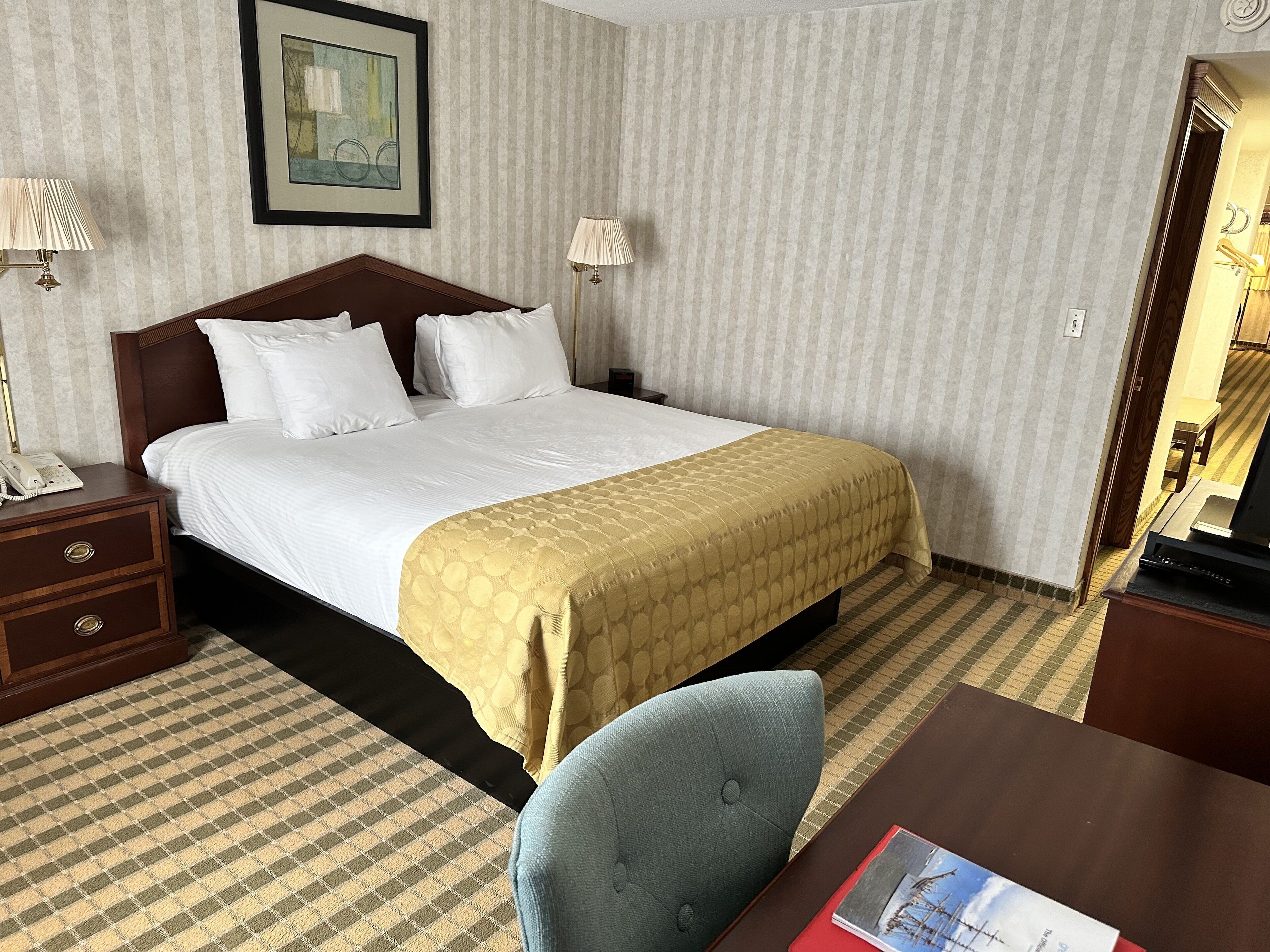 A hotel room with a large bed, a bedside table with a lamp, a chair, and a desk with a book.