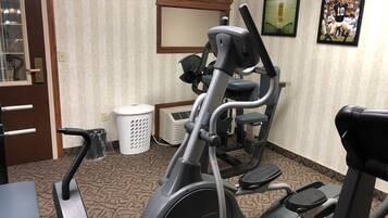 A fitness area with an elliptical machine, a stationary bike, and a treadmill. There is a door with a mirror, a white trash bin, and a framed picture on the wall.