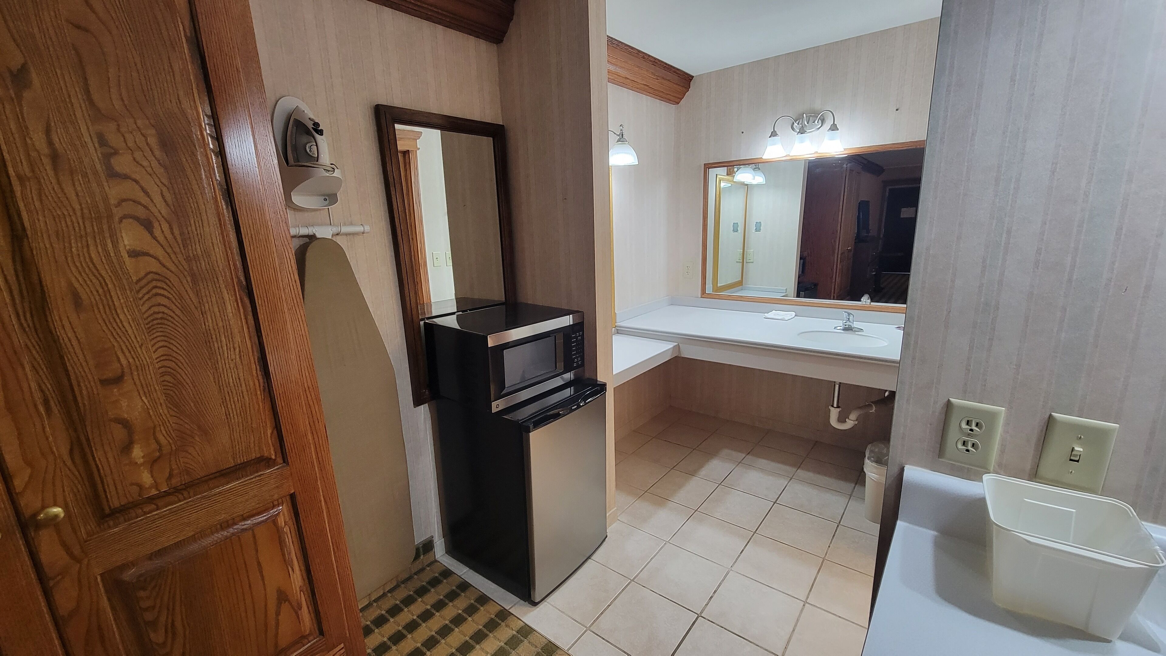 A compact bathroom with a wooden door, a built-in microwave, a sink with a mirror, and a trash bin.