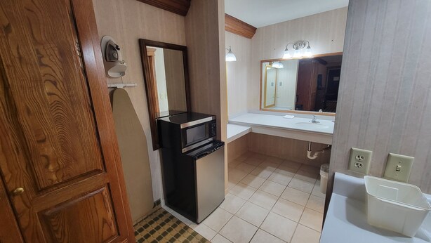 A compact bathroom with a wooden door, a built-in microwave, a sink with a mirror, and a trash bin. - Ramada by Wyndham Saginaw Hotel & Suites (Saginaw)