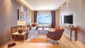 A modern living room with a sofa, coffee table, and armchair, a flat-screen TV, and a large window with a view.