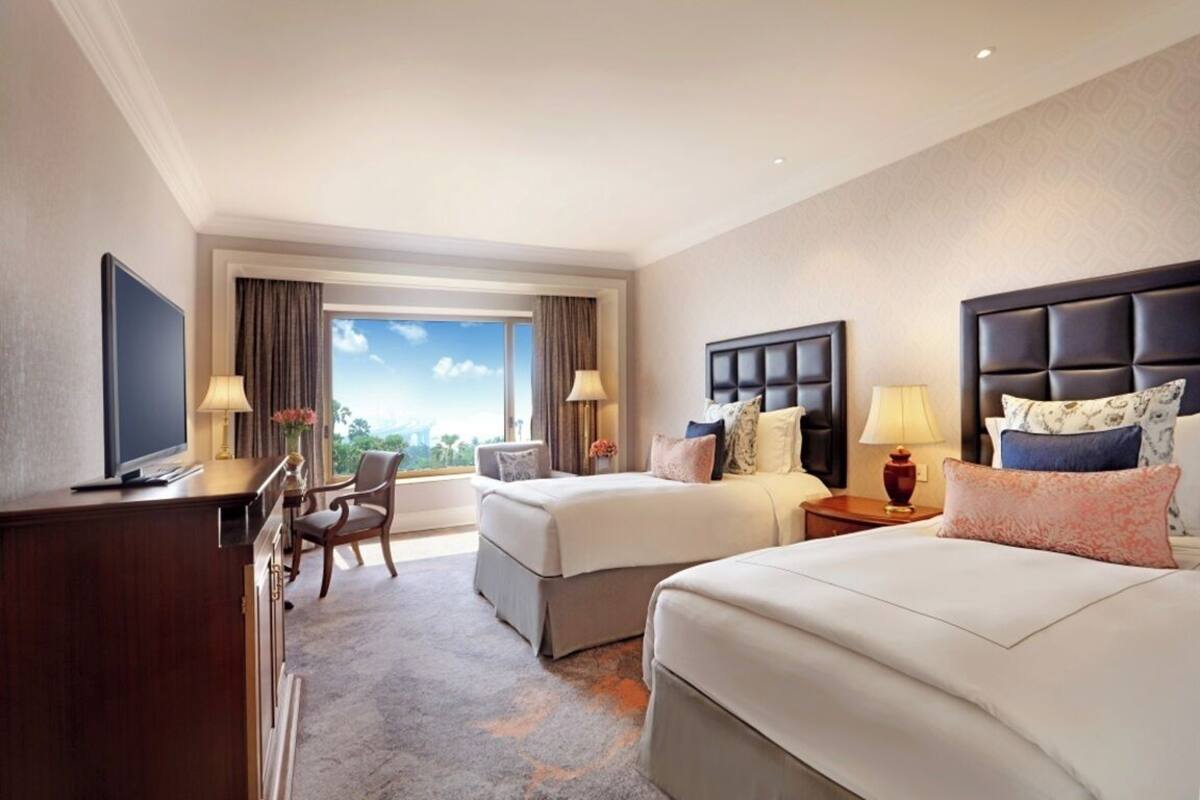 A hotel room with two beds, a desk, a TV, and a view of the sky.