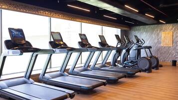 A row of treadmills and elliptical machines in a gym with wooden flooring and large windows.
