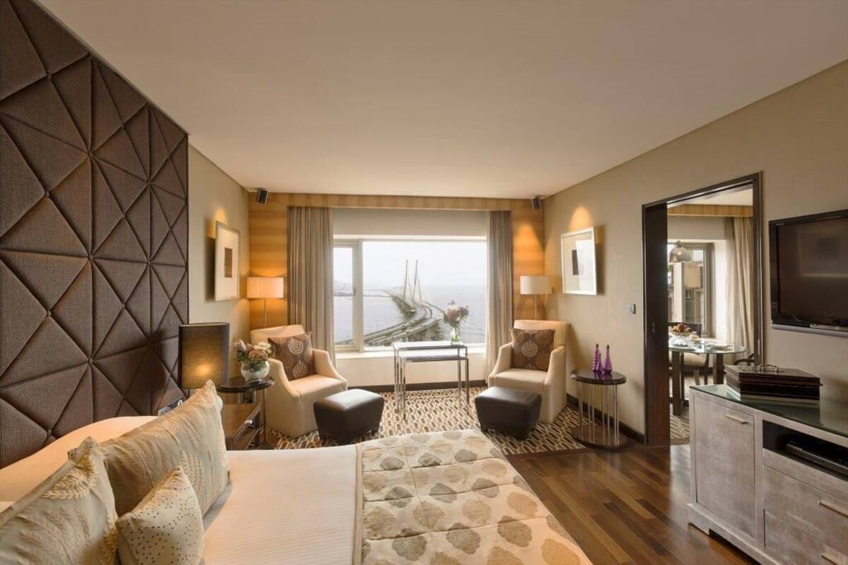 A hotel room with a large bed, two armchairs, a small table, a TV, and a view of a bridge.