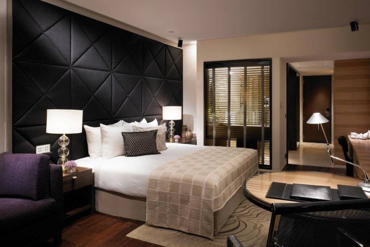A modern hotel room with a large bed, two bedside tables with lamps, a chair, a desk with a chair, and a large window with blinds.