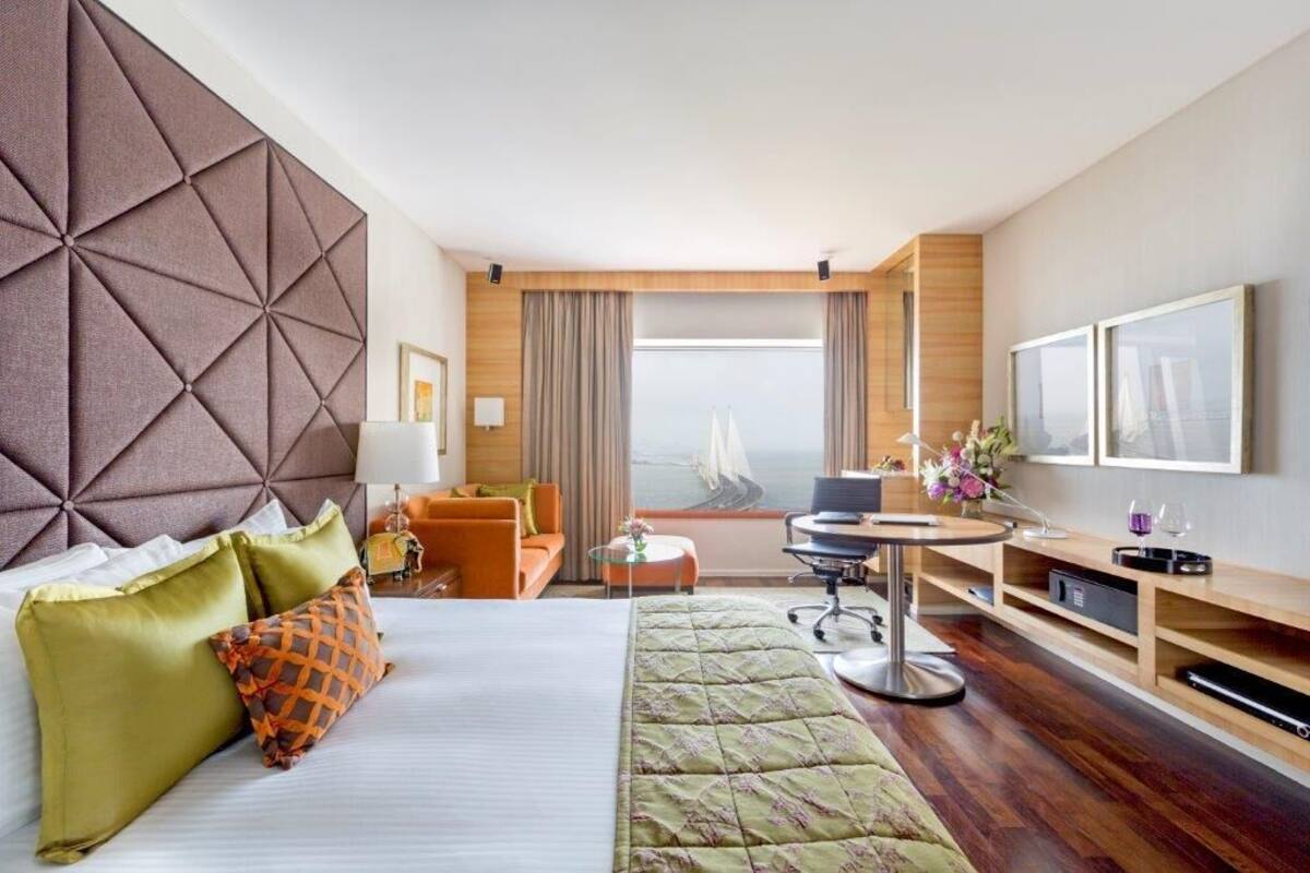 A modern hotel room with a large bed, a desk, a chair, a TV, and a view of the city.