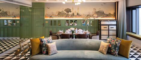 A hotel lobby with a checkered floor, a green reception desk, a dining area with a table set for two, and a sofa with floral cushions.