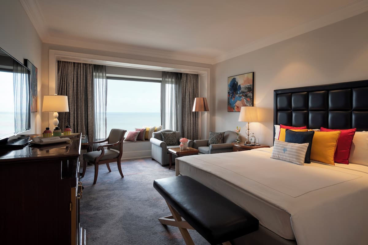 A hotel room with a large bed, a desk, a chair, and a sofa. There is a window with curtains, a painting on the wall, and a lamp on the desk.