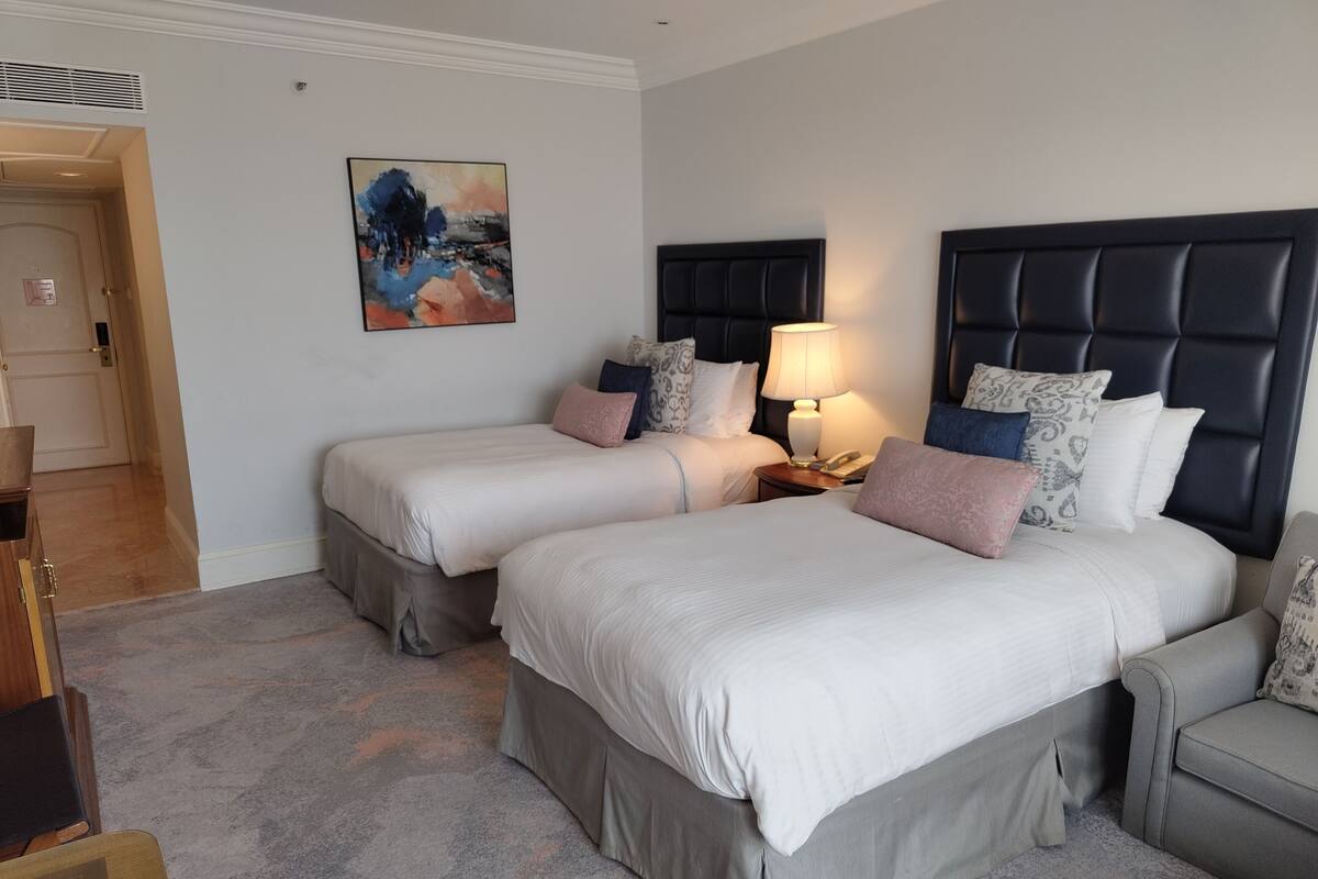 A hotel room with two beds, a nightstand with a lamp, a painting on the wall, and a door leading to another room.