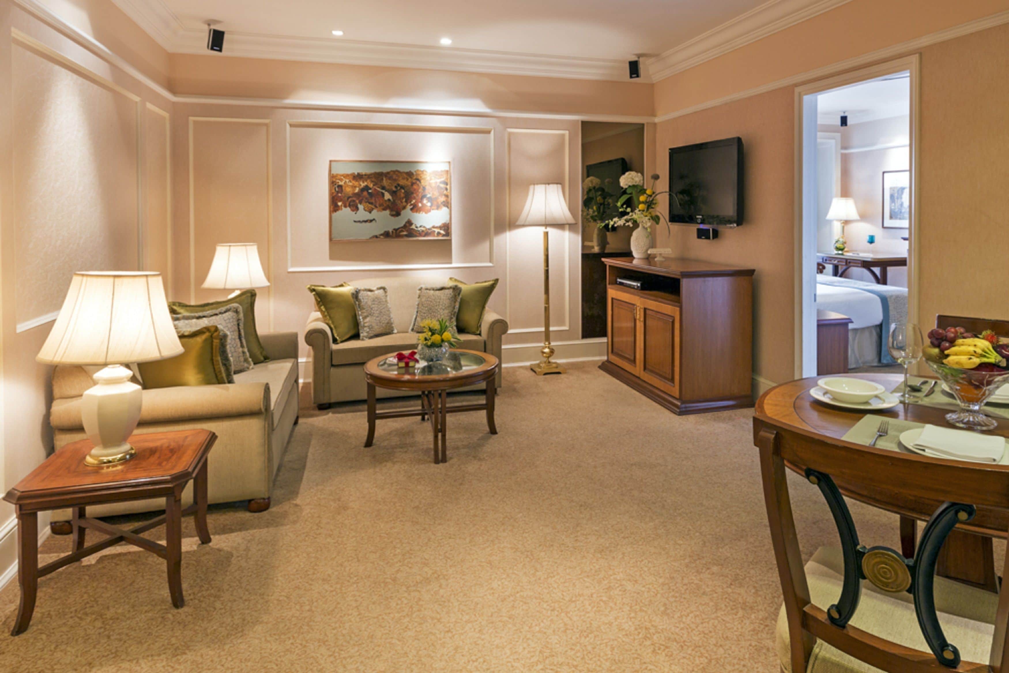 A well-lit living room with a sofa, coffee table, and lamps. There is a dining area with a table set for two and a television mounted on the wall.