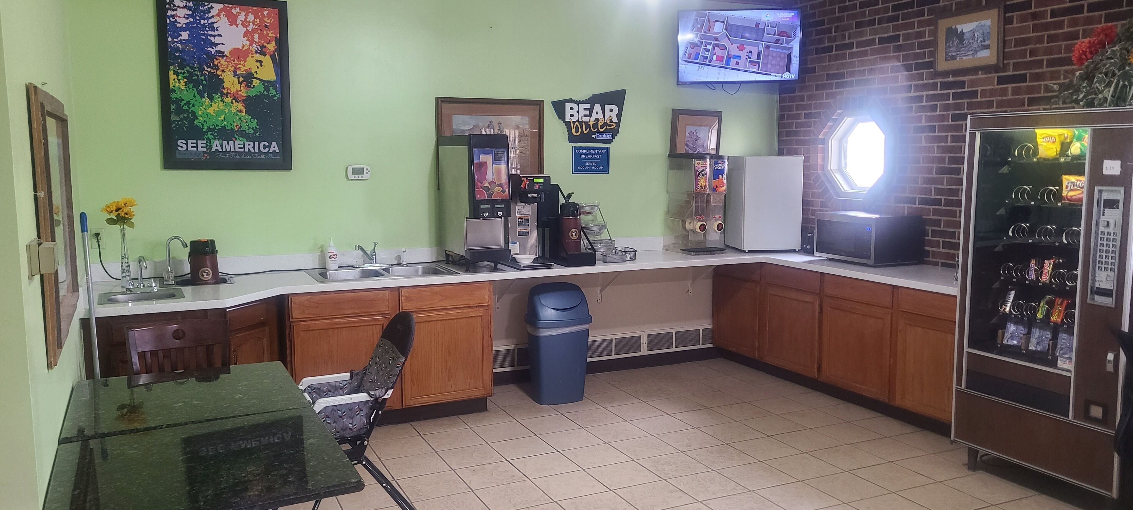 Uma cozinha com cafeteira, geladeira, lixeira e televisão fixada na parede.