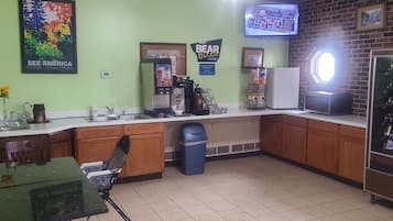 Uma cozinha com cafeteira, geladeira, lixeira e televisão fixada na parede.