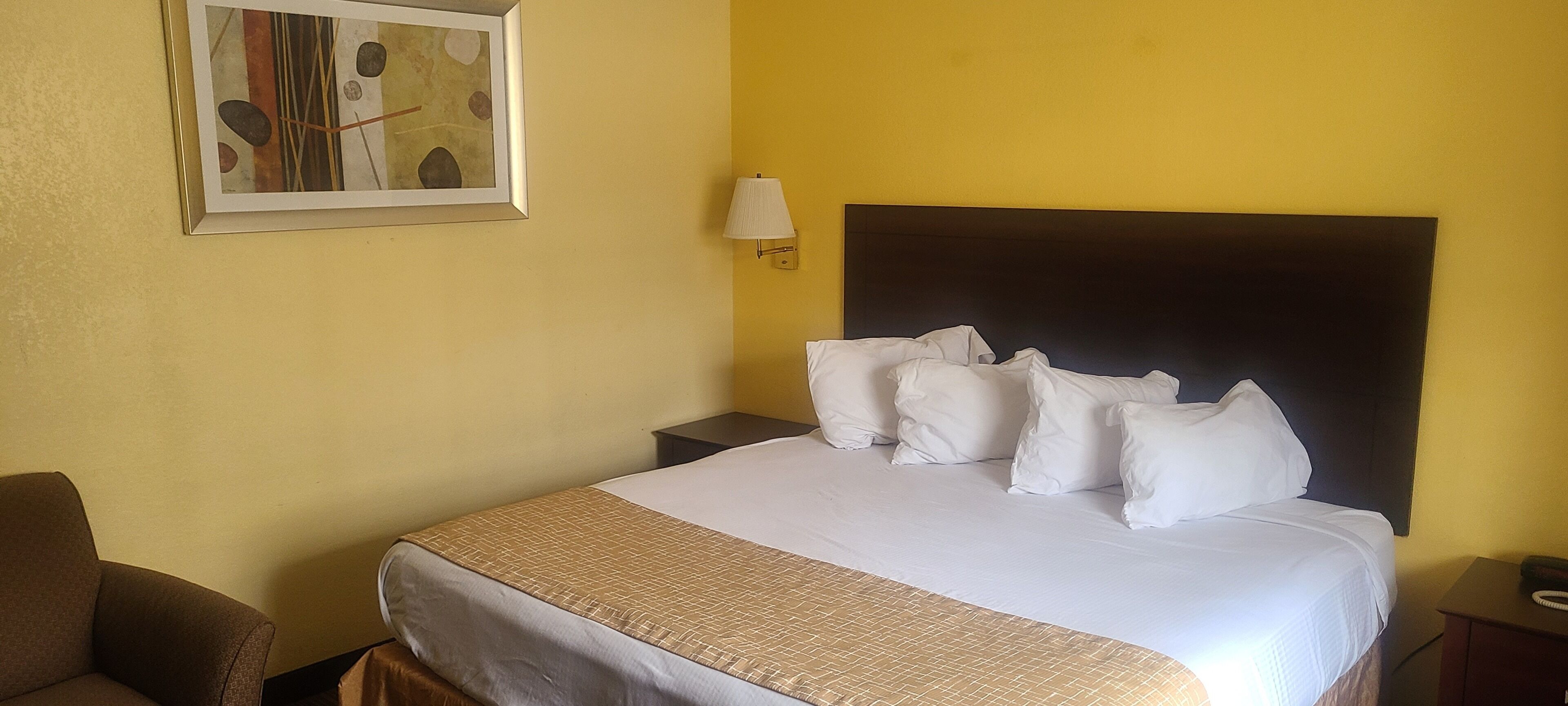 Una habitación de hotel con cama, mesita de noche, lámpara, silla y un cuadro en la pared.