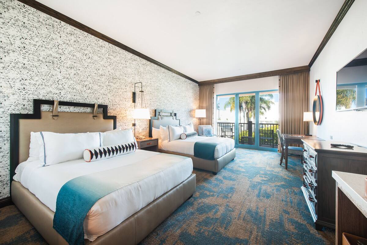 A hotel room with two beds, a desk, a chair, and a balcony with a view.