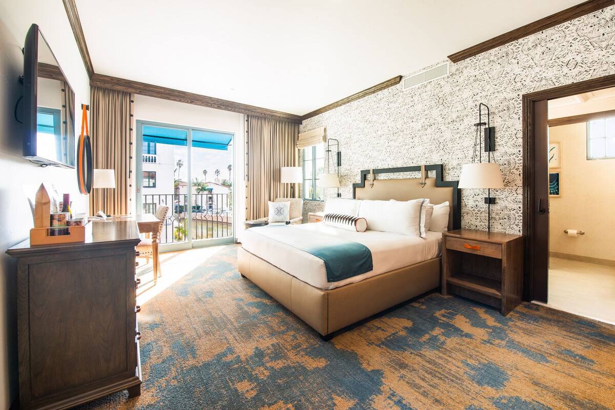 A hotel room with a large bed, a wooden dresser, a nightstand, and a balcony with a view.