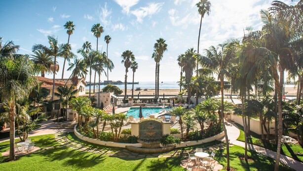 A resort with a swimming pool surrounded by palm trees, a beach view, and a well-maintained lawn. - Harbor View Inn (Santa Barbara)