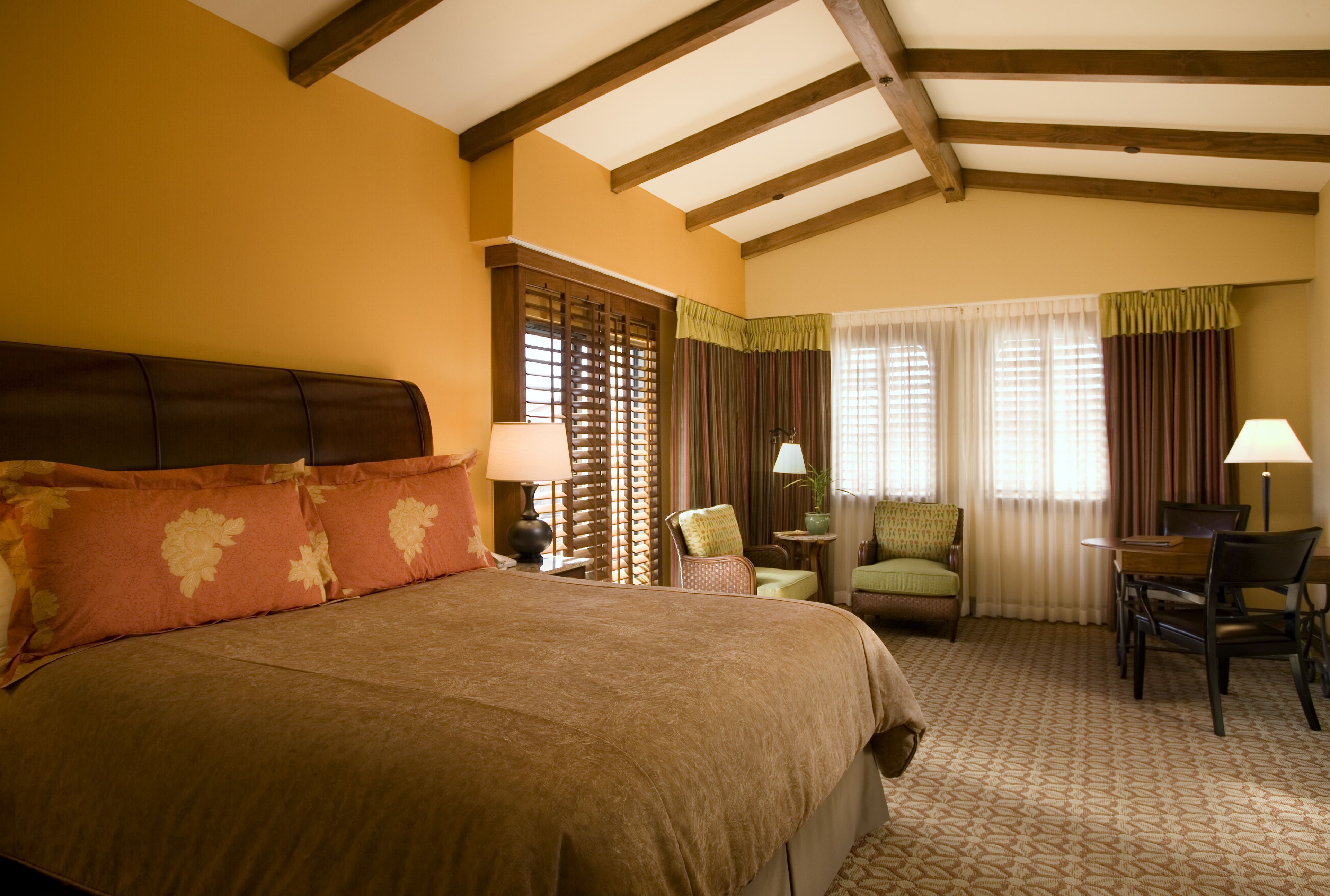 A hotel room with a large bed, a desk, two chairs, a window with curtains, and a wooden ceiling.