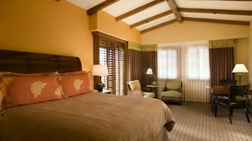 A hotel room with a large bed, a desk, two chairs, a window with curtains, and a wooden ceiling.