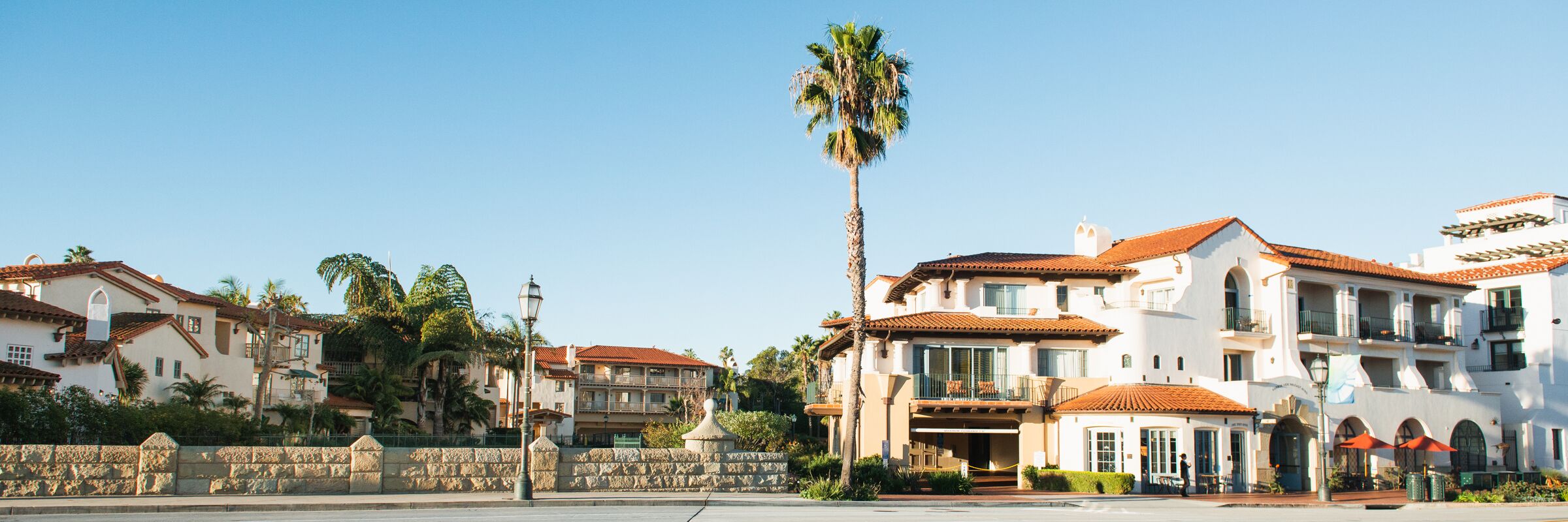 A coastal hotel with Mediterranean architecture, featuring multiple balconies, a stone wall, and palm trees.