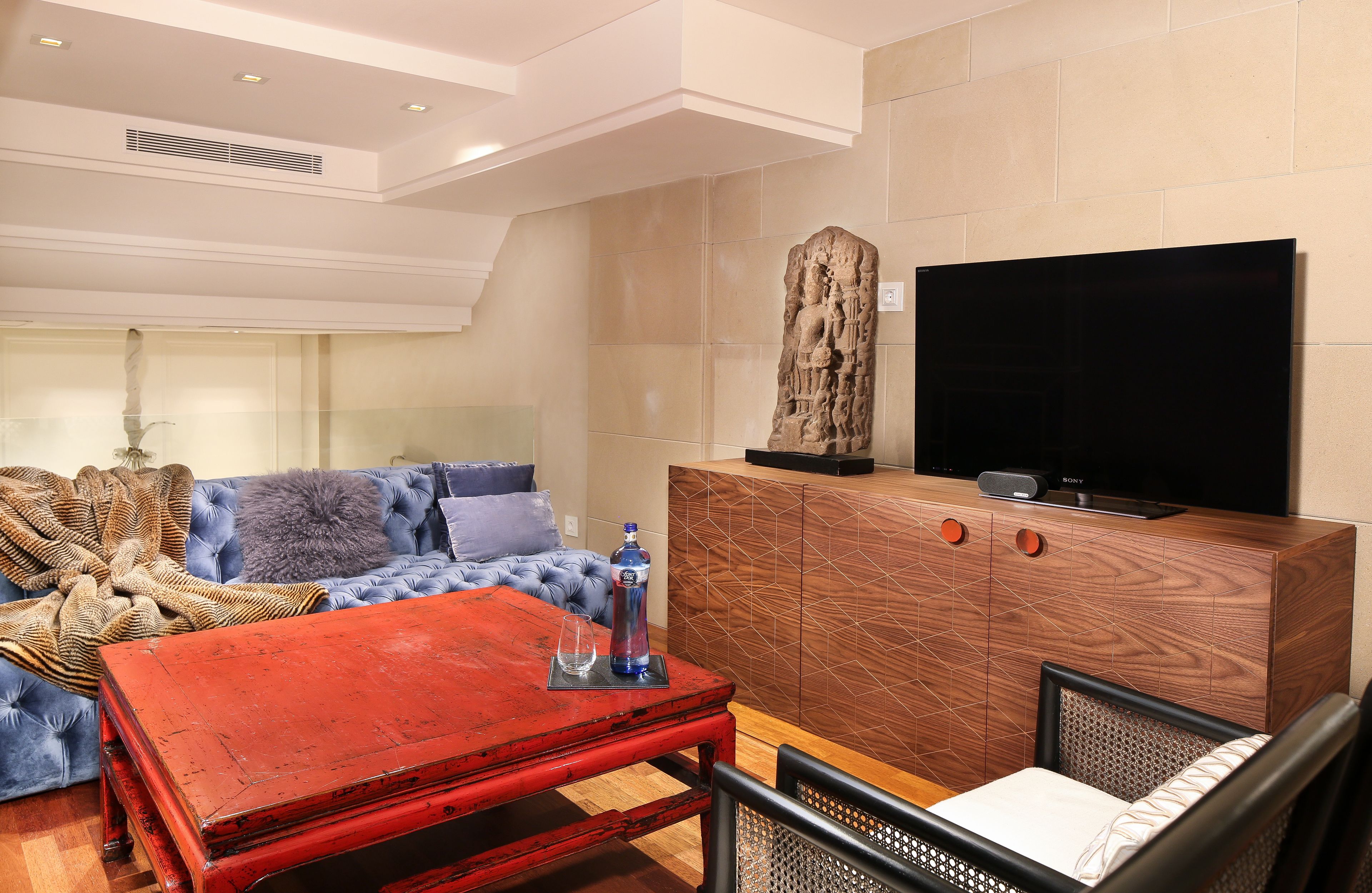 Een moderne woonkamer met een rode salontafel, een blauwe bank, een houten kast met een flatscreen televisie en een decoratief stenen beeldhouwwerk.