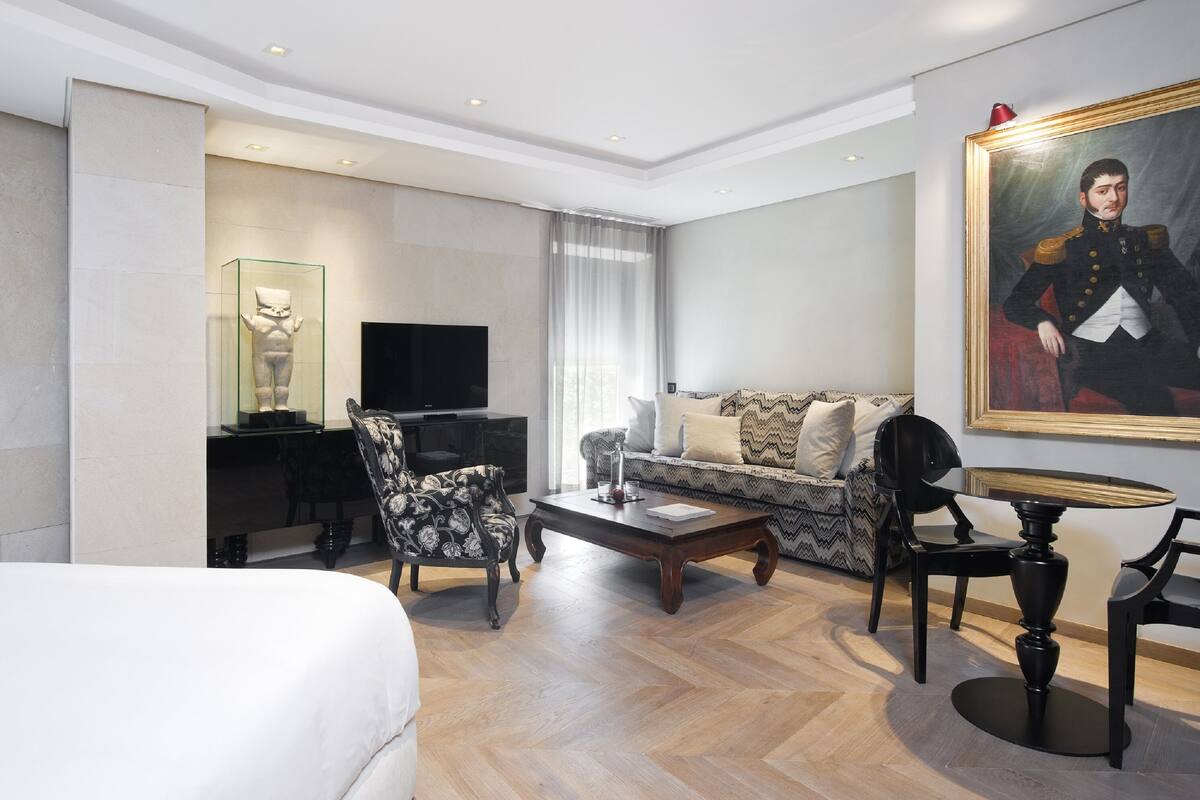 A modern hotel room with a large bed, a sofa, a dining table, a TV, and a painting on the wall.