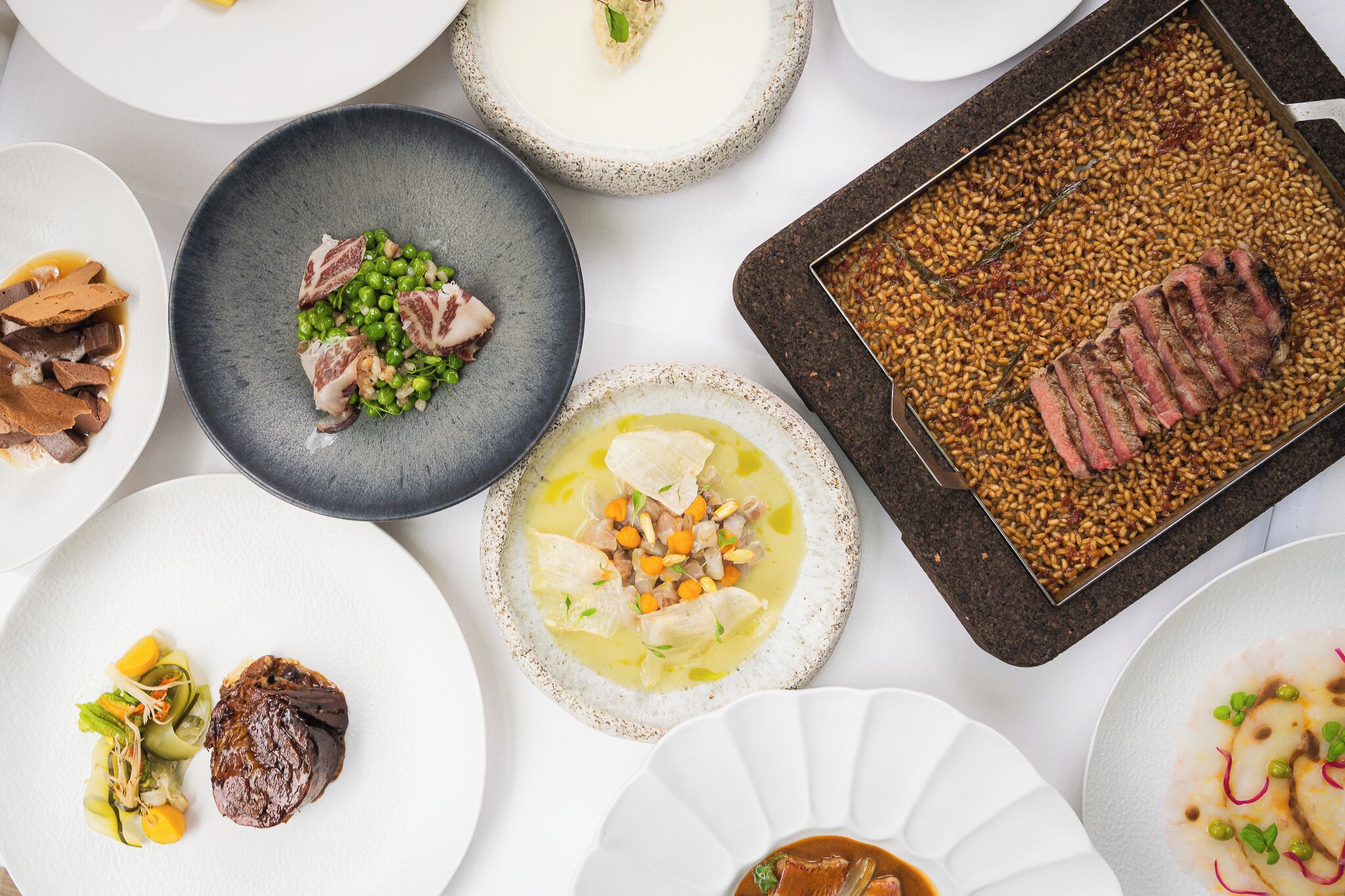 A variety of dishes on white plates, including a dish with meat, peas, and carrots, and another with a steak and beans.