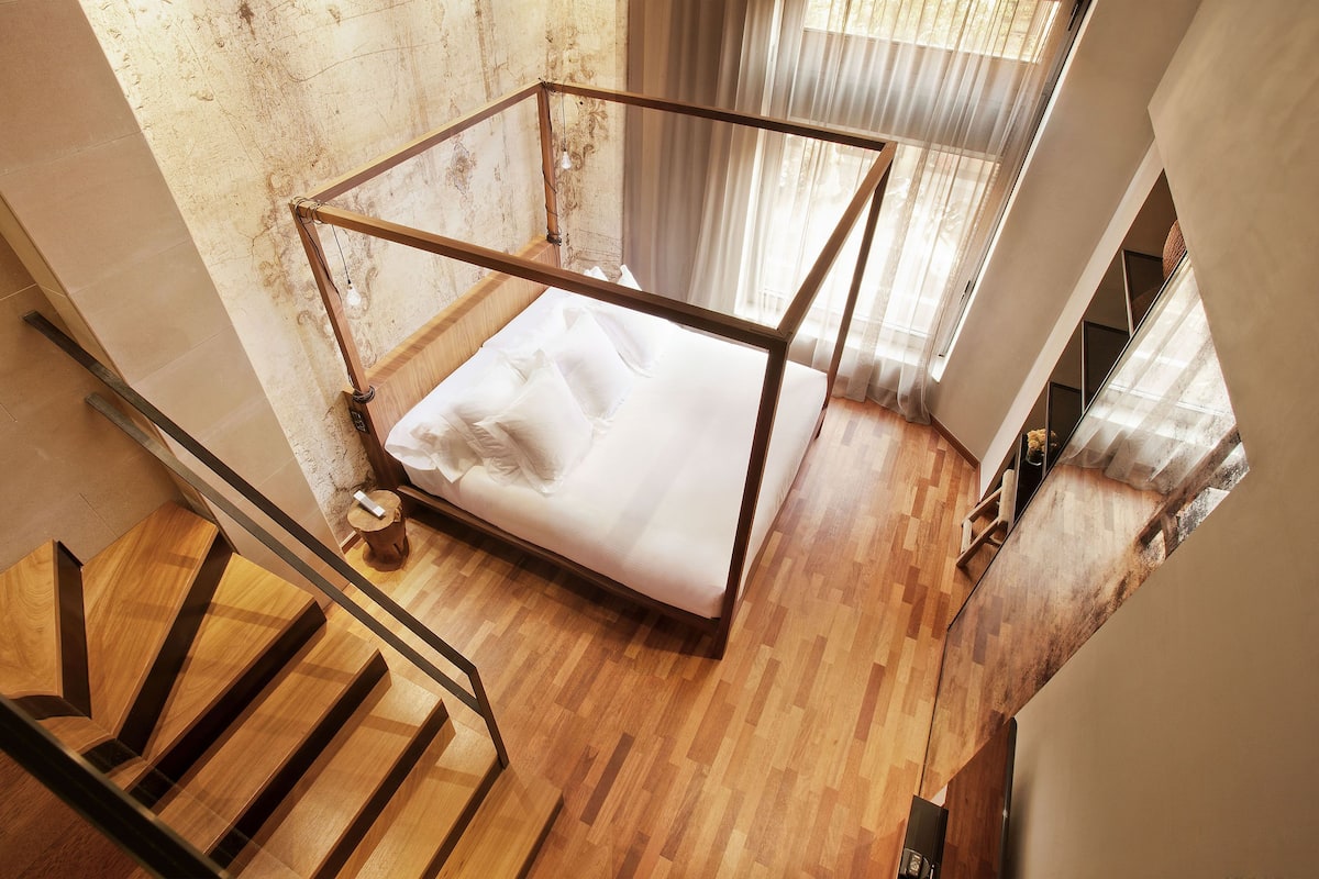 A high-ceilinged room with a wooden floor, a metal bed frame, and a staircase.