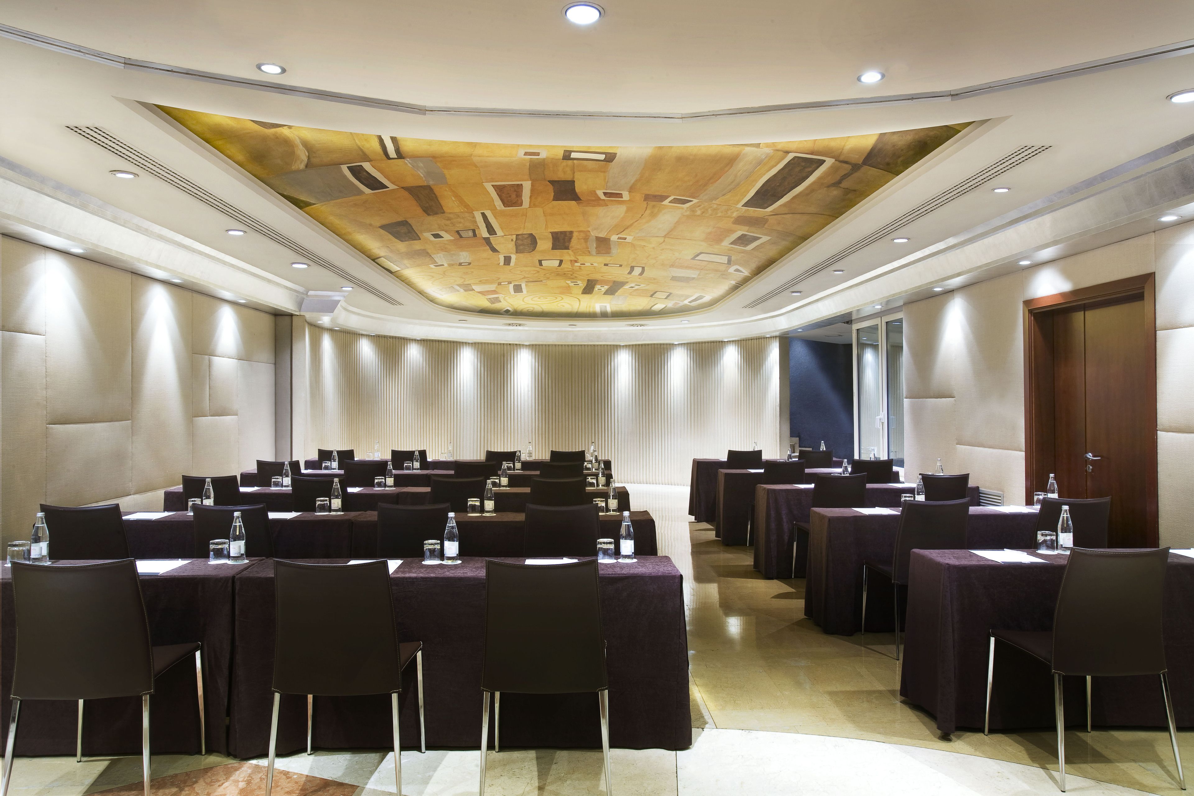 Une salle de conférence avec des tables et des chaises, un plafond orné de motifs et une grande porte sur le côté droit.