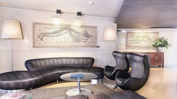 Una sala de estar moderna con un sofá de cuero negro curvo, una mesa de centro de vidrio y dos sillones negros. Hay dos lámparas de pared grandes y un tapiz de gran tamaño.