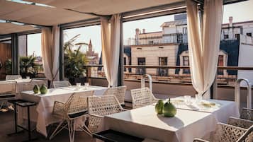 Een restaurant met witte tafels en rieten stoelen, grote ramen met gordijnen en uitzicht op gebouwen.