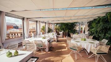 Un área de comedor al aire libre con mesas, sillas, ventanas amplias y plantas en macetas.