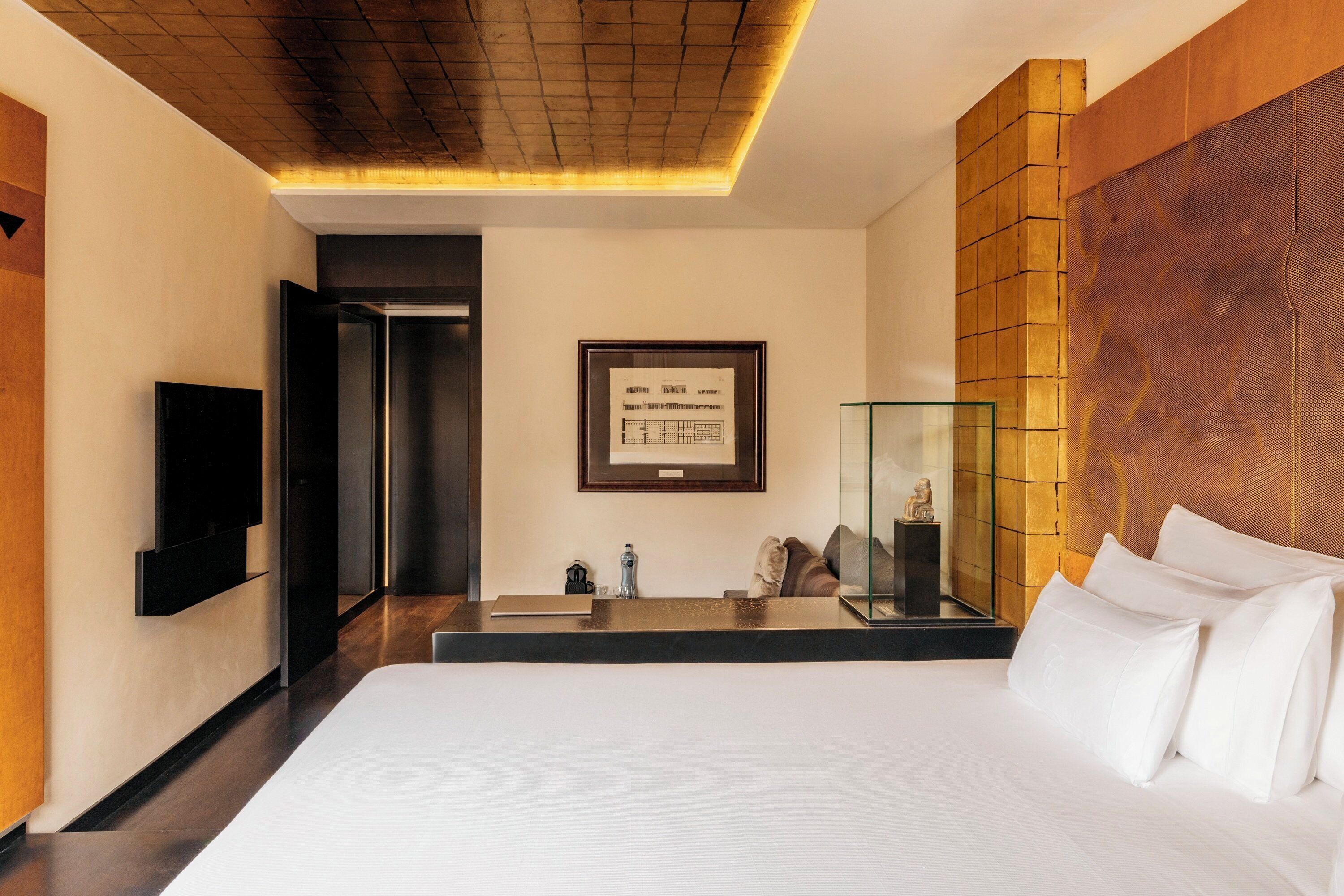 A modern hotel room with a large bed, a flat-screen TV mounted on the wall, a framed architectural drawing, and a glass display case.