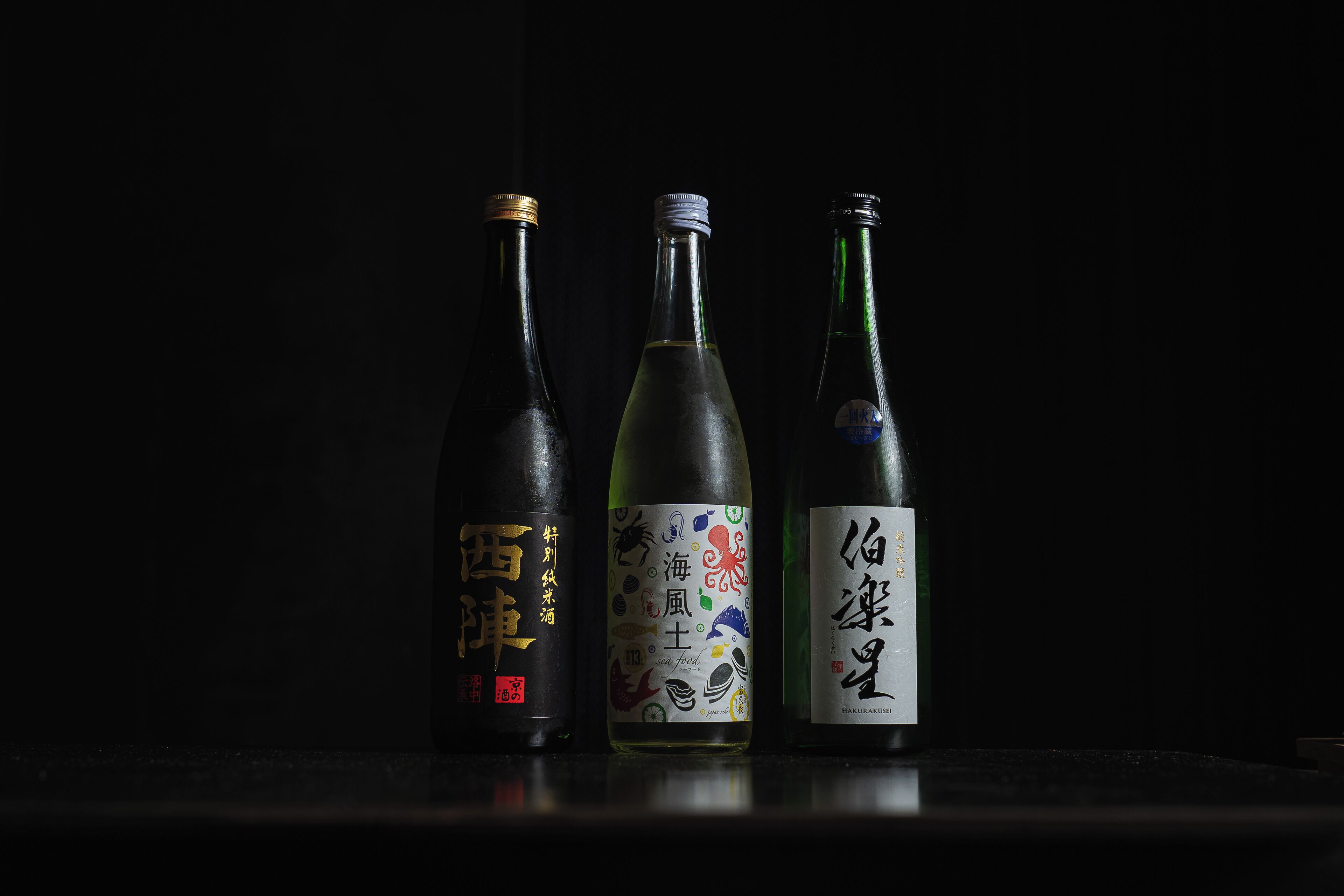 Tres botellas de sake sobre una superficie reflectante, sobre un fondo oscuro.