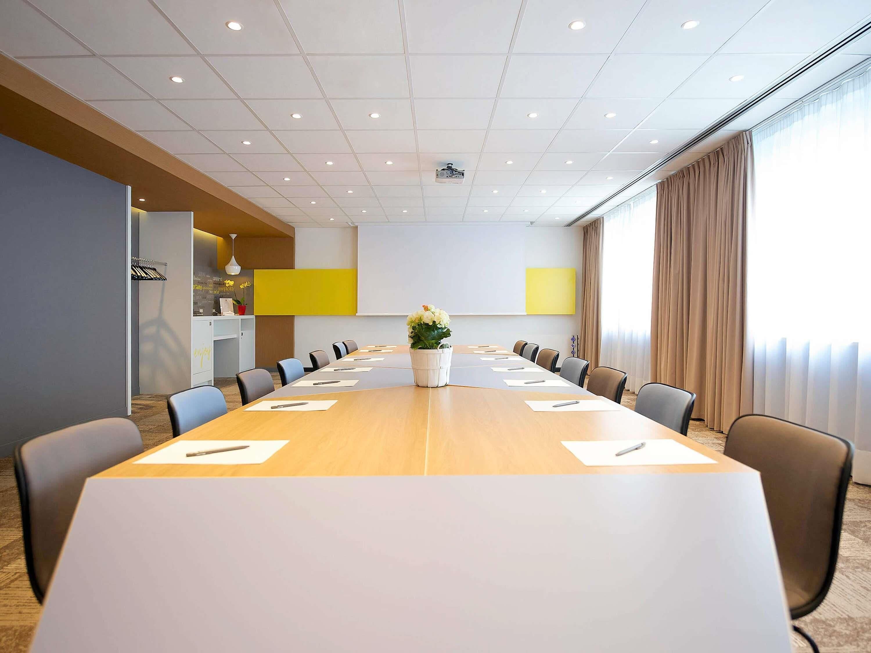 Un salón de conferencias con una larga mesa, sillas, una pantalla de proyector y un amplio ventanal con cortinas.