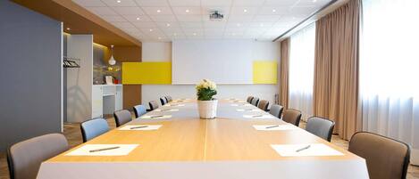 Ein Konferenzraum mit langem Tisch, Stühlen, einer Projektionsleinwand und einem großen Fenster mit Vorhängen.