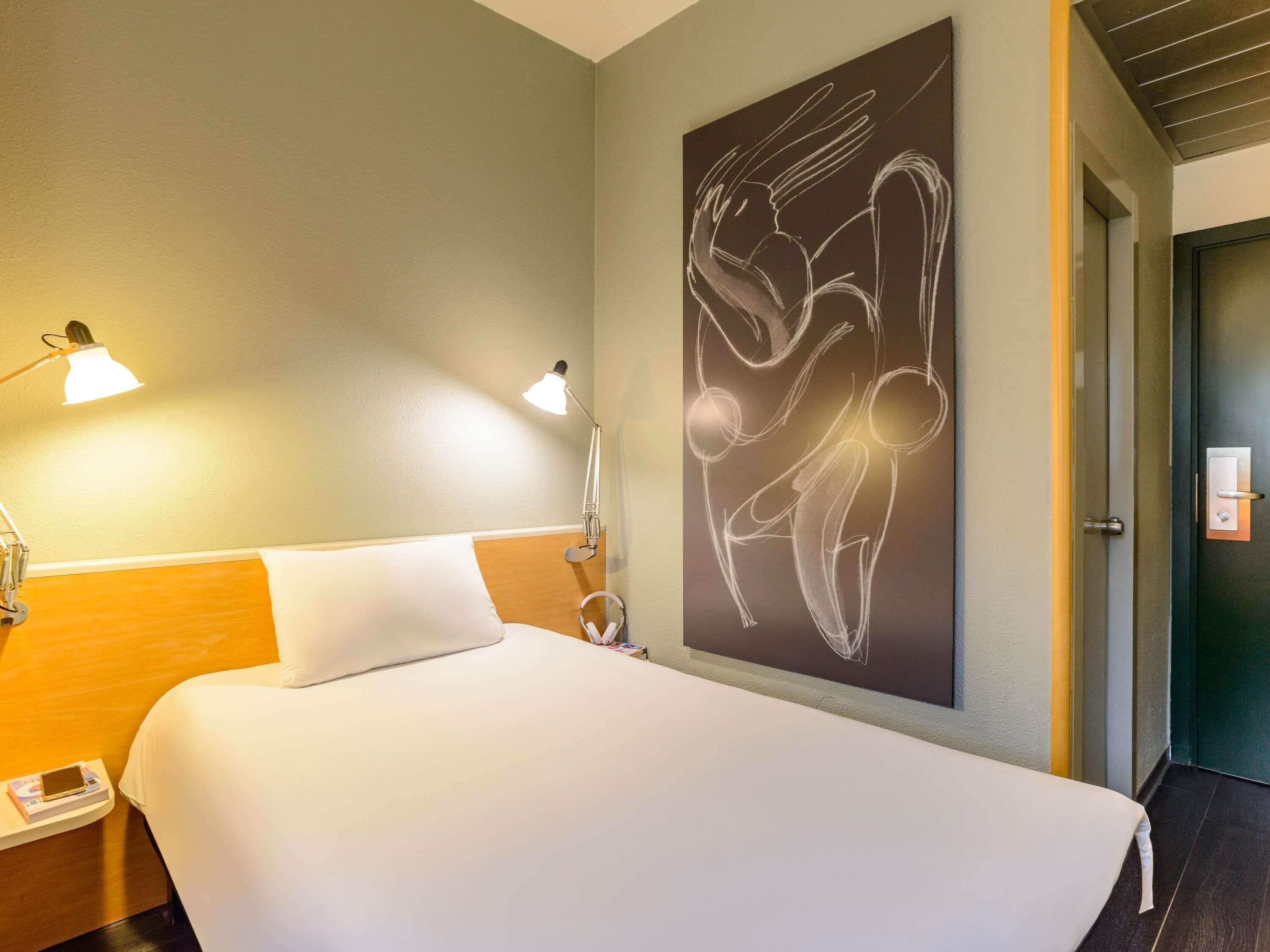 A hotel room with a large bed, two bedside lamps, a wall-mounted abstract artwork, and a door to the right.