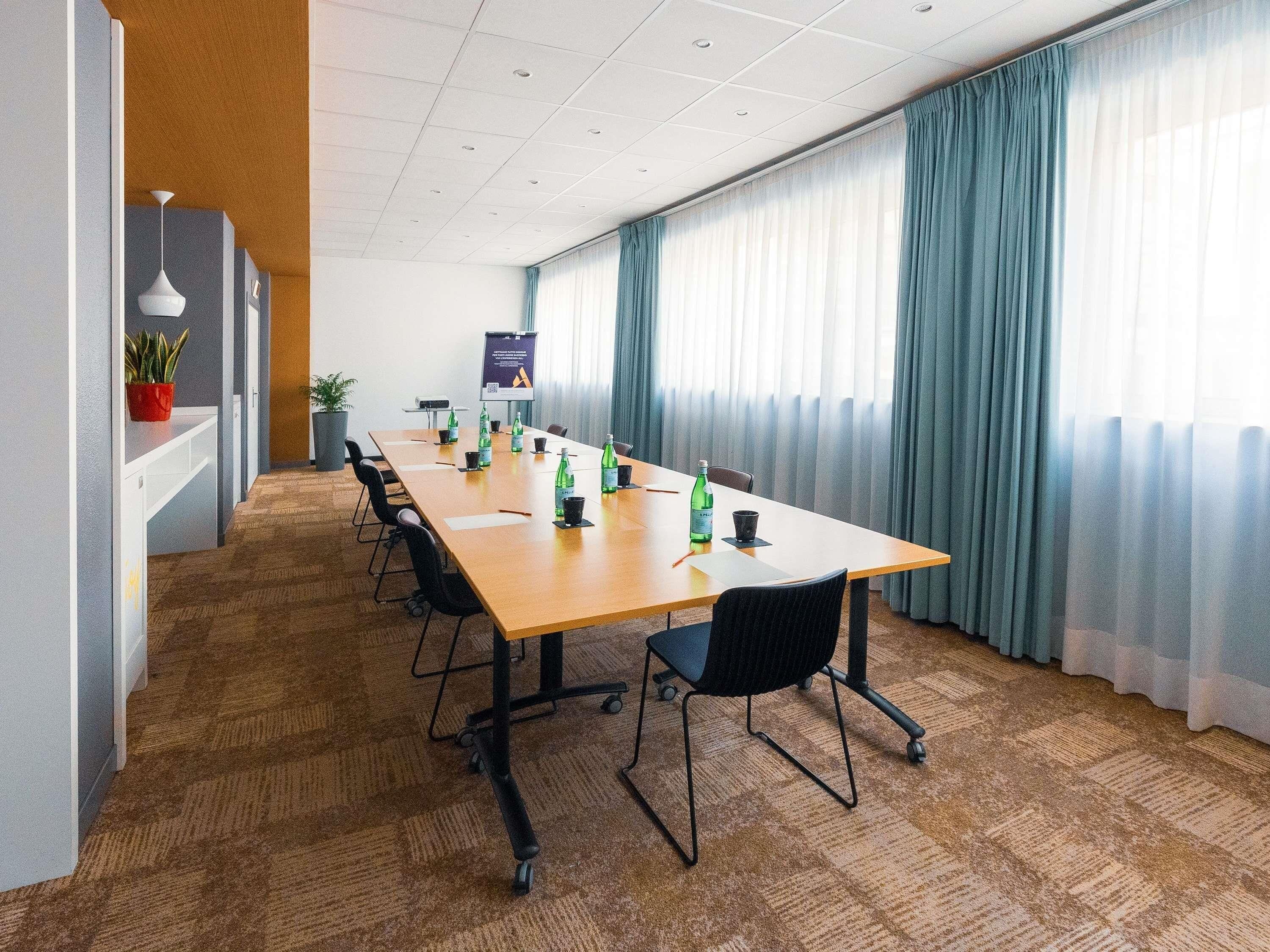 Ein Konferenzraum mit einem langen Tisch, Stühlen, einer Projektionsleinwand und einem großen Fenster mit Vorhängen.