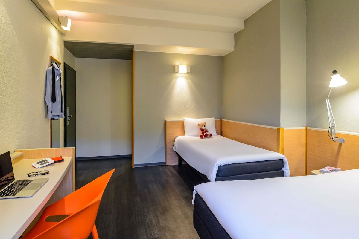 A hotel room with a desk, two beds, a chair, a lamp, and a teddy bear on one of the beds.