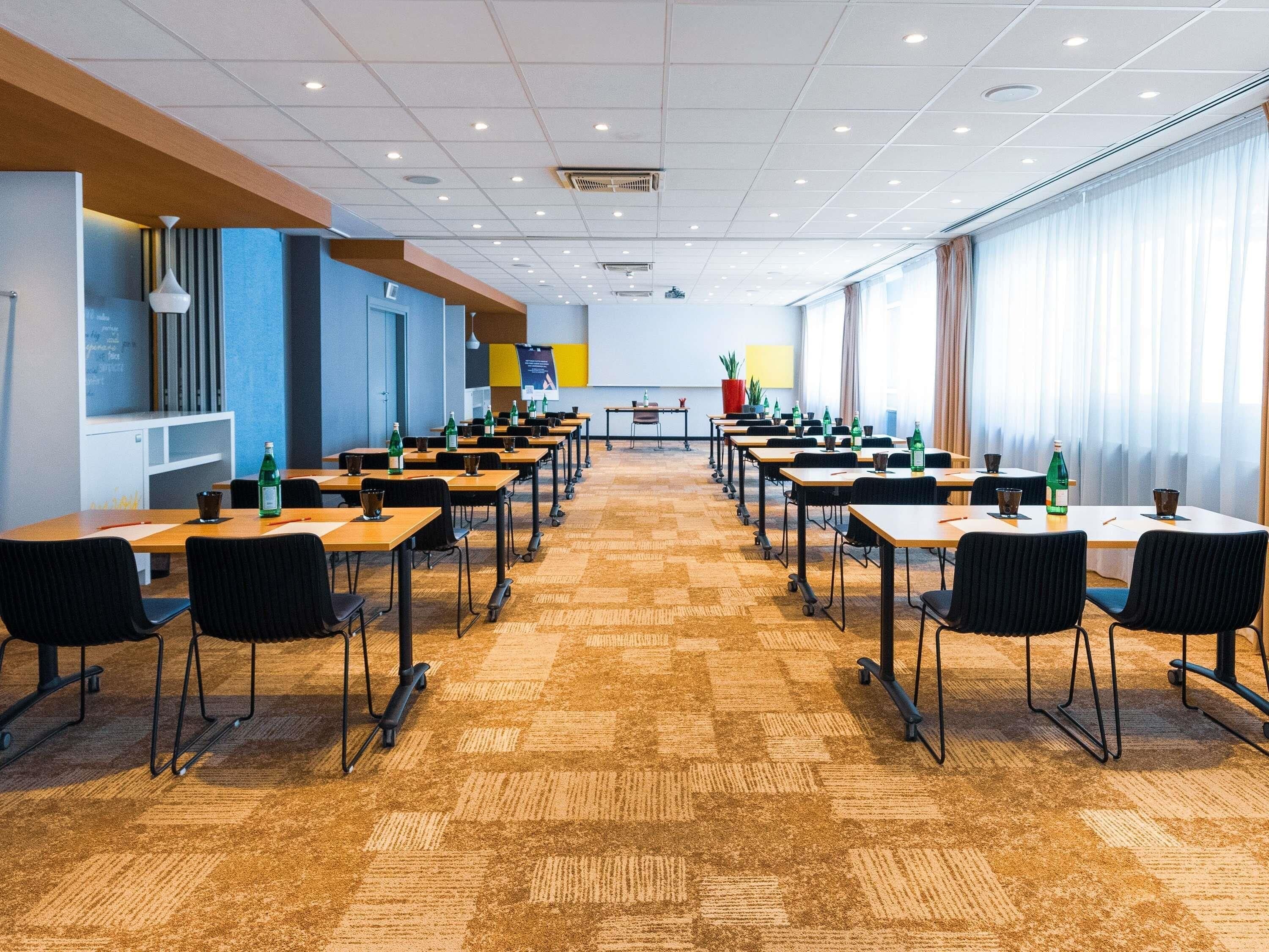 Une salle de conférence dotée de plusieurs tables et chaises, d’un sol recouvert de moquette et de grandes fenêtres avec des rideaux.