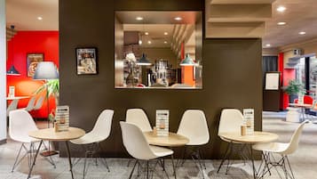 Un moderno hall de hotel con una zona de estar central, un espejo grande que refleja el interior, y un mostrador de restaurante visible al fondo.