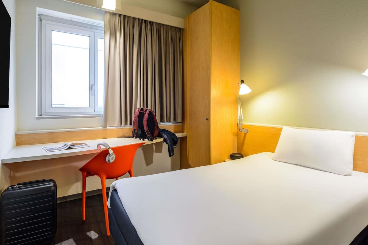Una habitación de hotel con una cama, un escritorio con una silla, una maleta, una lámpara y una ventana con cortinas.