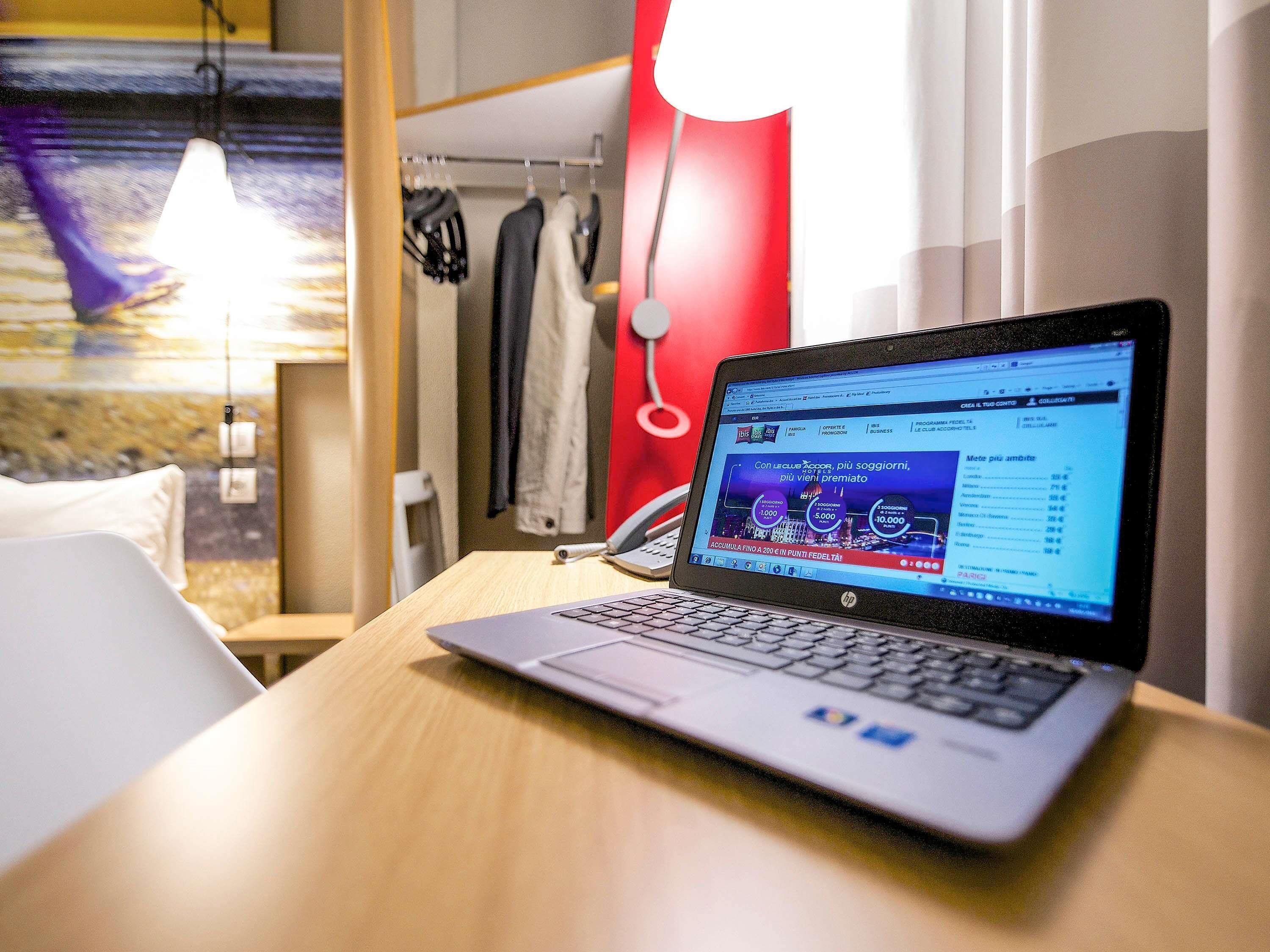 Ein Laptop auf einem Holztisch in einem Hotelzimmer, mit einem Bett, einer roten Wand und einer Pendelleuchte.
