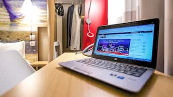 Ein Laptop auf einem Holztisch in einem Hotelzimmer, mit einem Bett, einer roten Wand und einer Pendelleuchte.