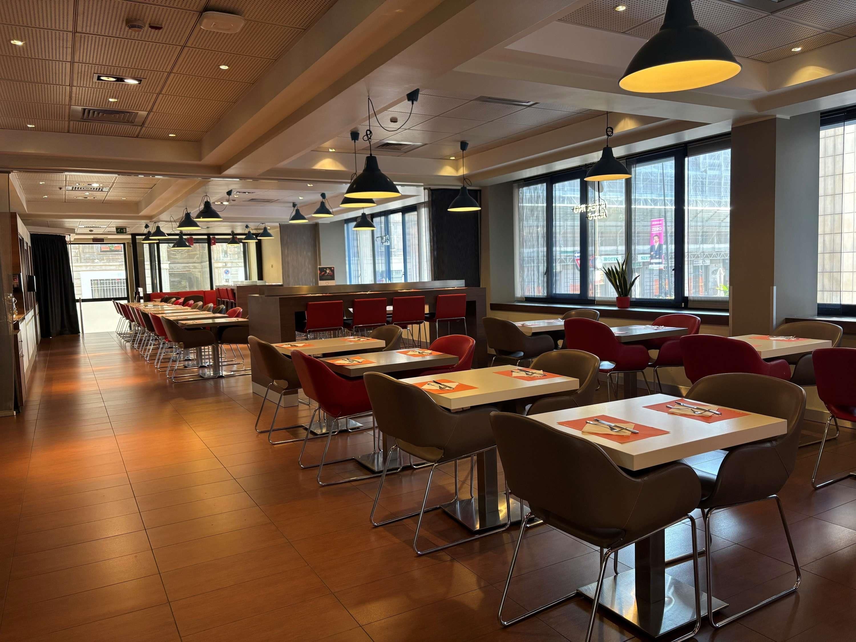 Un intérieur de restaurant moderne, avec de nombreuses tables et chaises, de grandes fenêtres et des luminaires suspendus.