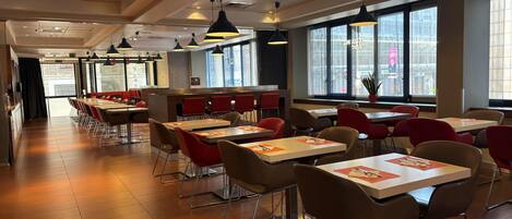 Un intérieur de restaurant moderne, avec de nombreuses tables et chaises, de grandes fenêtres et des luminaires suspendus.