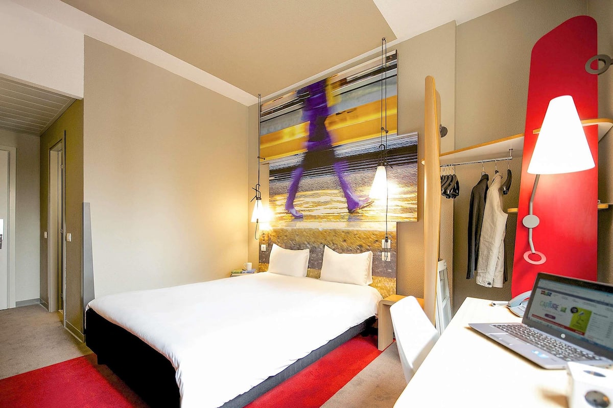 A hotel room with a bed, a desk with a laptop, a red carpet, and a wall with a colorful abstract painting.