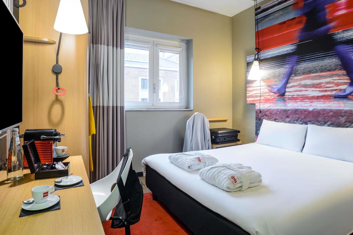 A hotel room with a bed, a desk with a coffee maker, a chair, a window, and a painting on the wall.