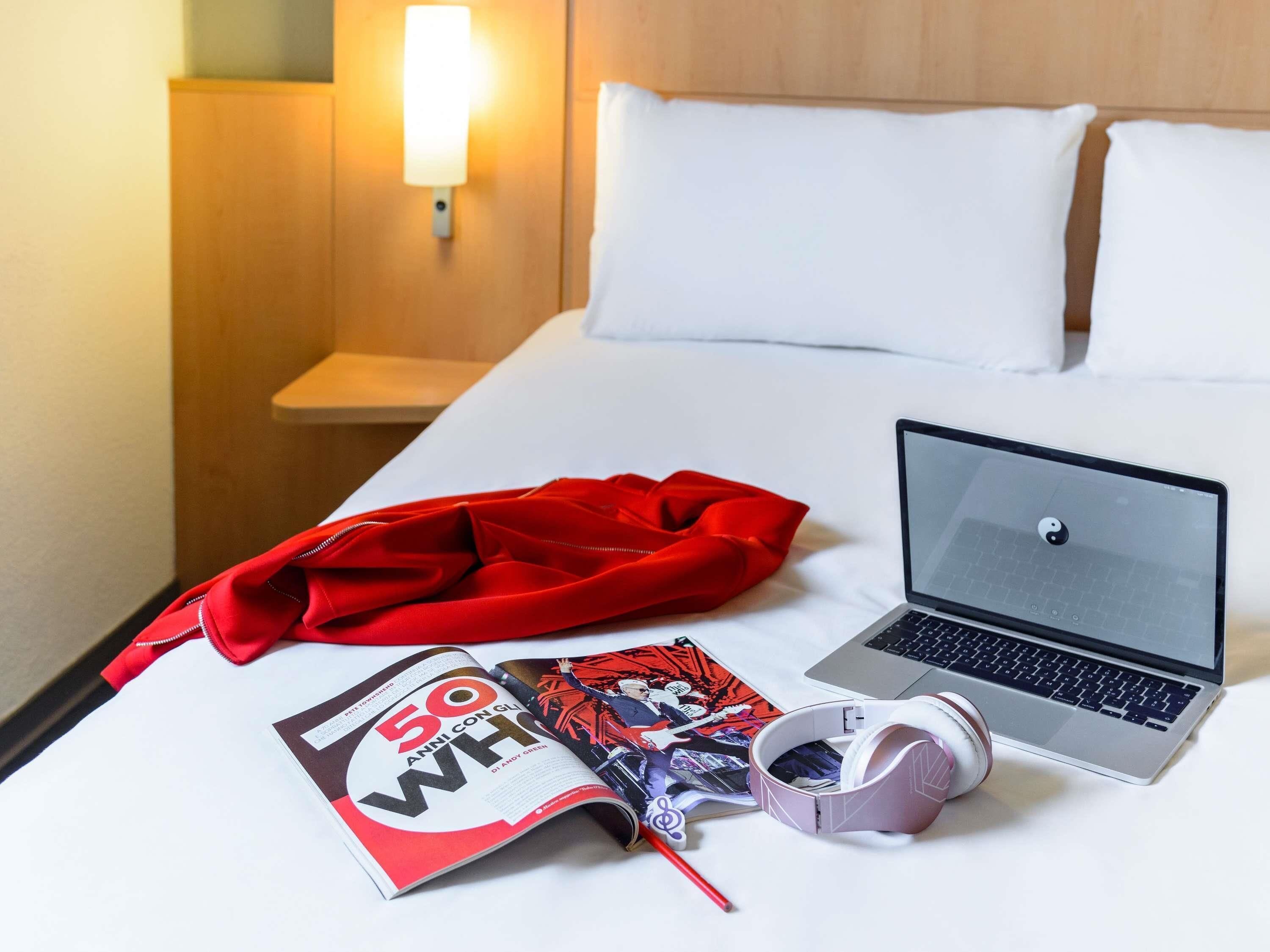 Hotellihuone, jossa on sänky, kannettava tietokone, punainen takki, lehti ja kuulokkeet.