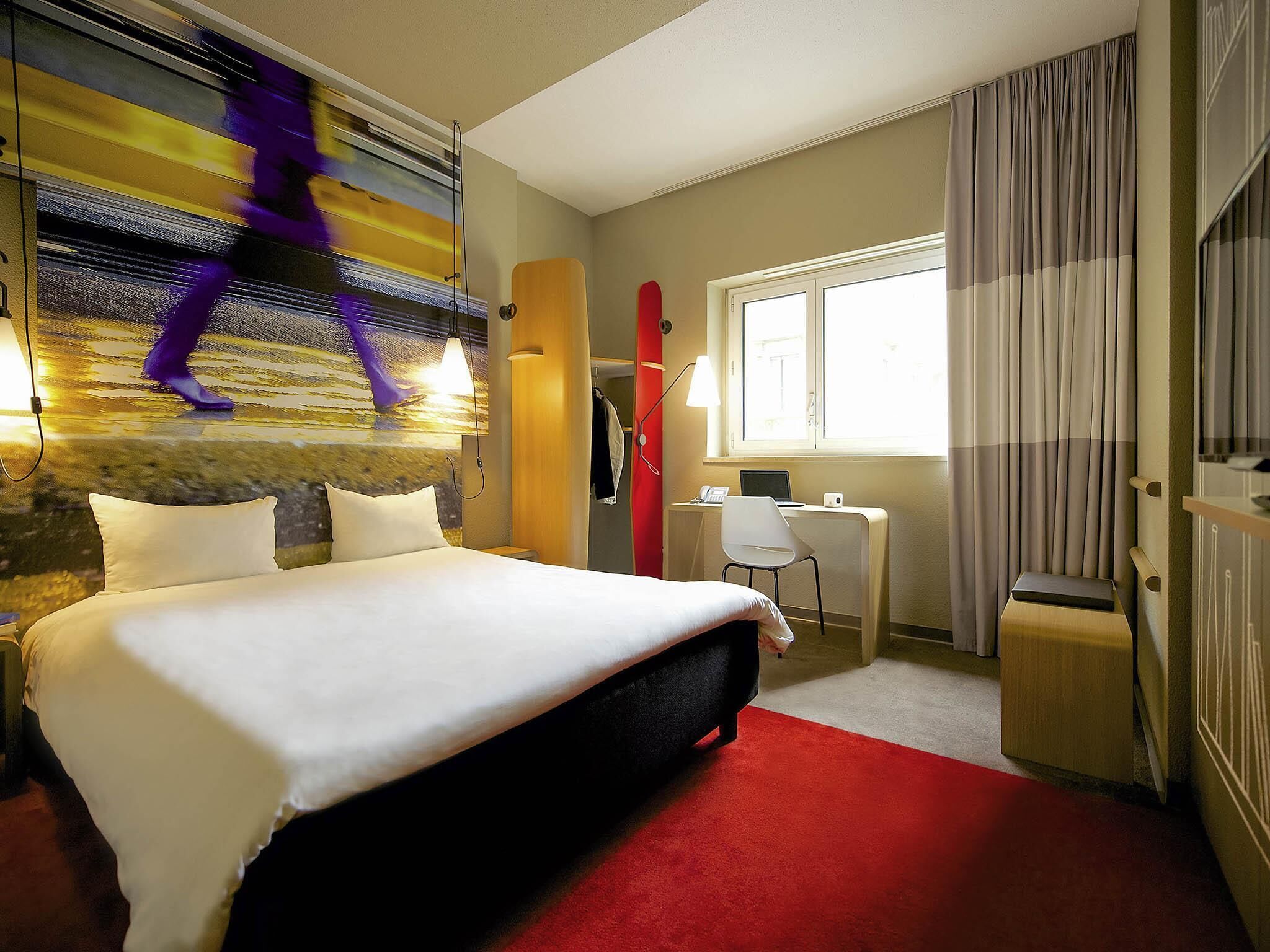 A hotel room with a bed, a desk, a chair, a window with curtains, and a wall with a mural.