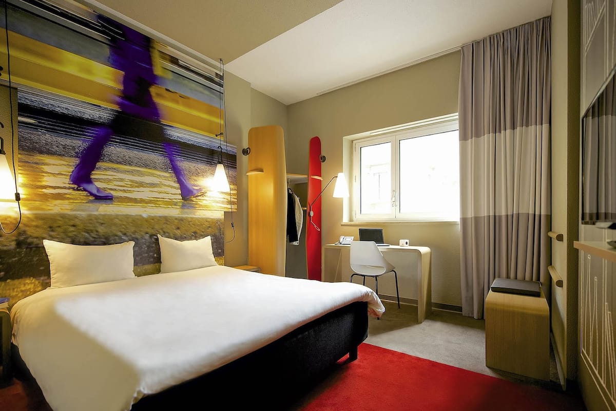 A hotel room with a bed, a desk, a chair, a window with curtains, and a wall with a mural.